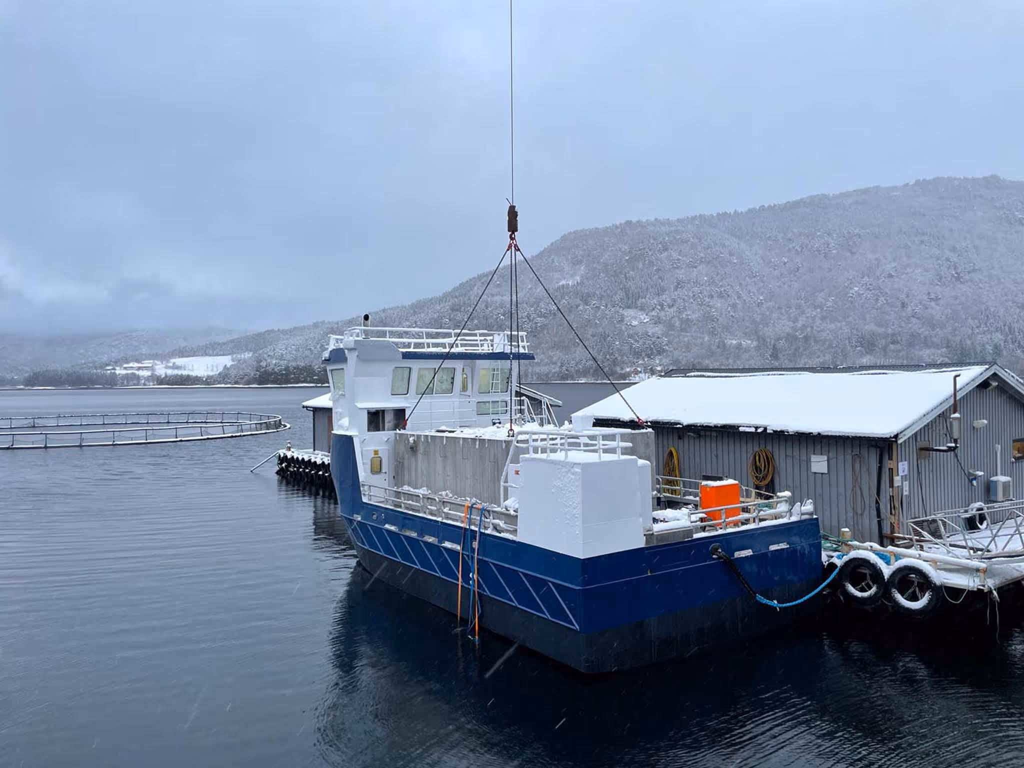 Blå og hvit båt bundet til kai ved en snødekket bygning med snødekte fjell i bakgrunnen.