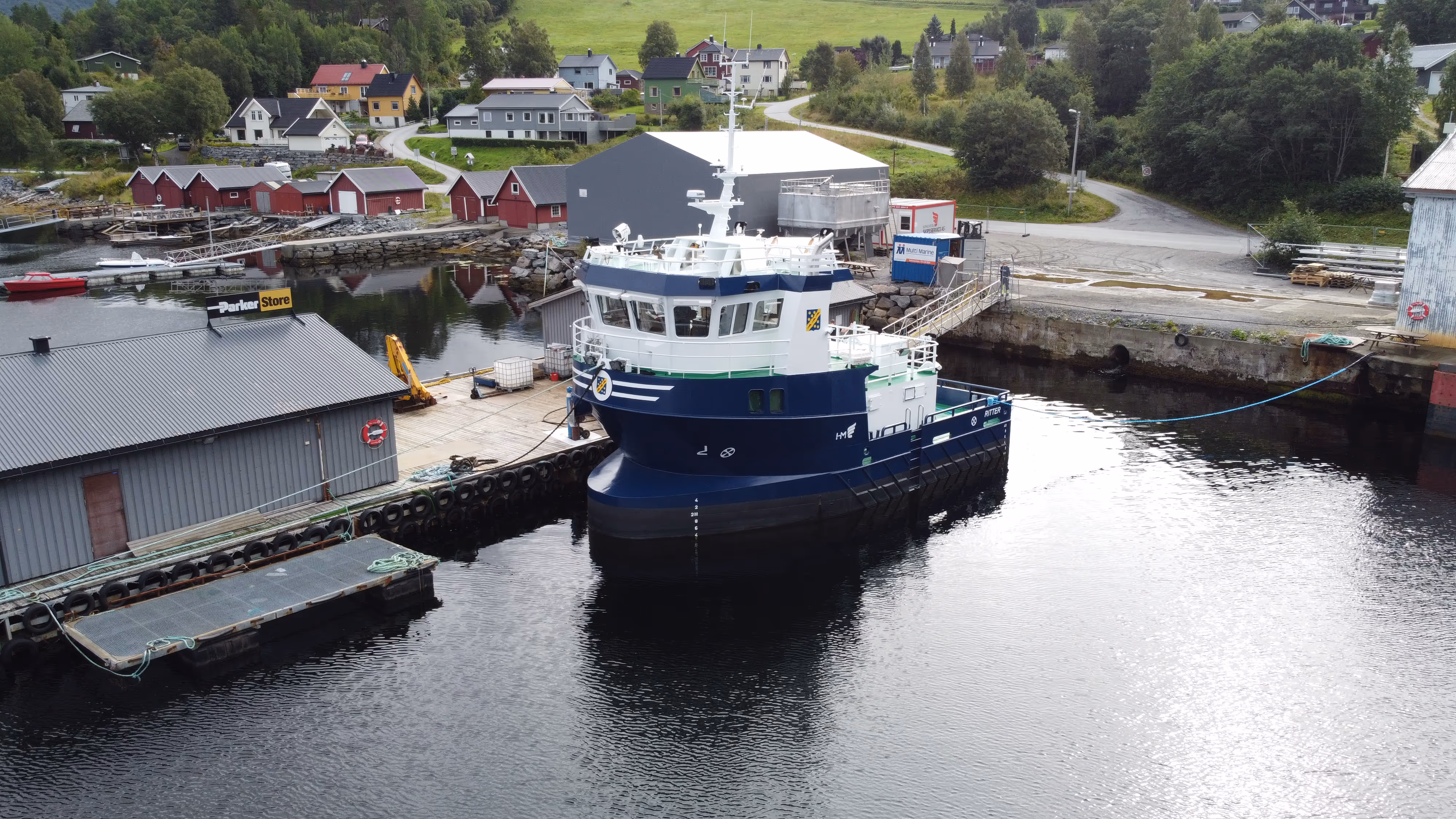 Blått og hvitt fartøy fortøyd ved kaien i en liten havn med bygninger og grønn vegetasjon i bakgrunnen.