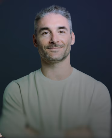 Smiling man with short gray hair and light facial stubble wearing a beige shirt against a dark blurred background.