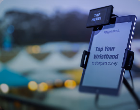 Electronic display screen with a black device above it showing 'TAP HERE!' and text on the screen reading 'Tap Your Wristband to Complete Survey', with a blurred background of outdoor tents and lights.