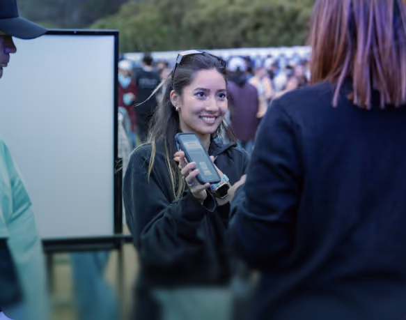 A smiling woman holding a smartphone and showing its screen to another person during an outdoor event with a crowd.