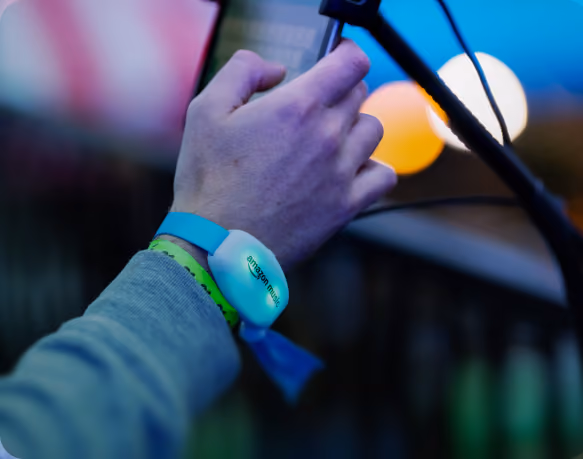 Close-up of a person’s hand holding a microphone with a wristband that has an Amazon Music tag, set against colorful background lights.