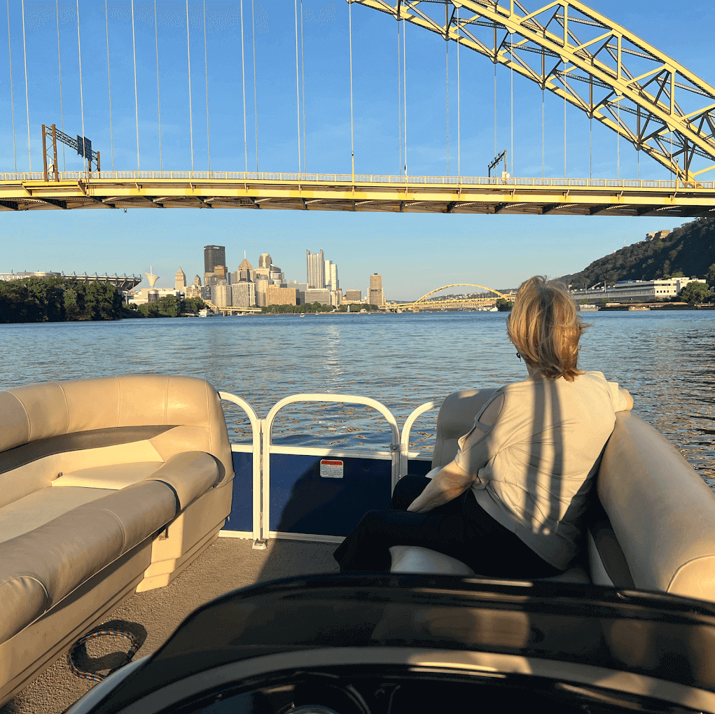 A woman seated at the front of a pontoon passing under the West End Bridge toward the downtown skyline in warm light