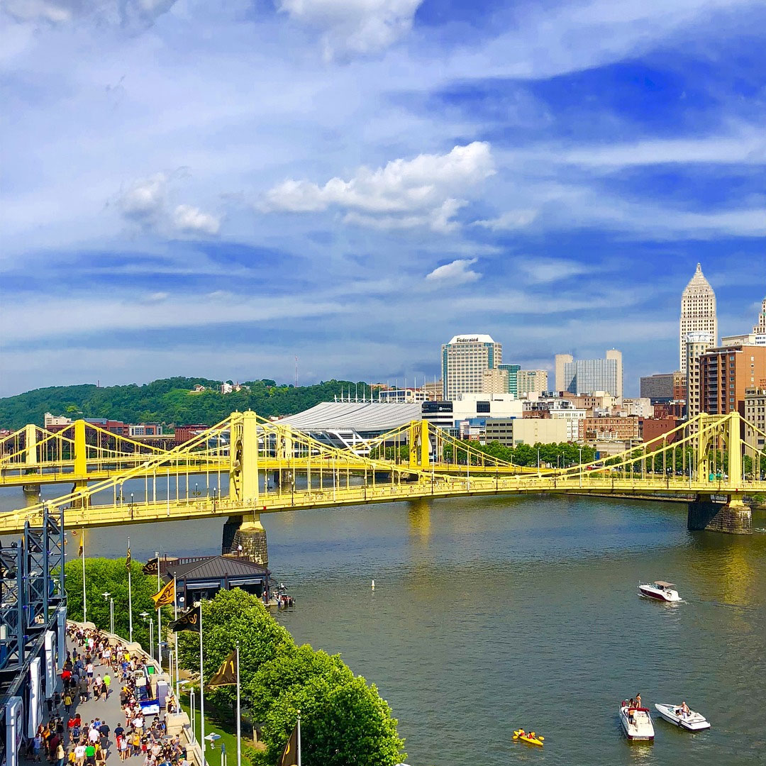 Elevated view of the yellow Roberto Clemente Bridge, downtown skyline and crowds on the riverwalk, with boats and a kayak on the river