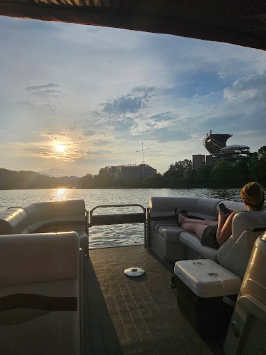 A woman lounging on a pontoon seat looking at her phone as the sun sets, with Acrisure Stadium and the Science Center on the shore.