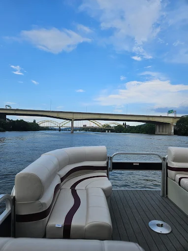 An empty pontoon deck with white and maroon seats heading toward a concrete highway bridge with yellow arch bridges beyond