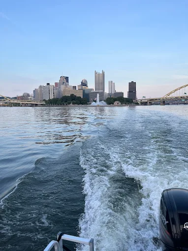 The white wake behind a pontoon's outboard motor, with the Point fountain and downtown skyline behind in evening light.