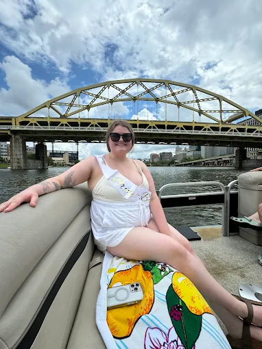 A smiling woman wearing a bride sash sitting on a pontoon seat with a yellow double deck arch bridge behind her.