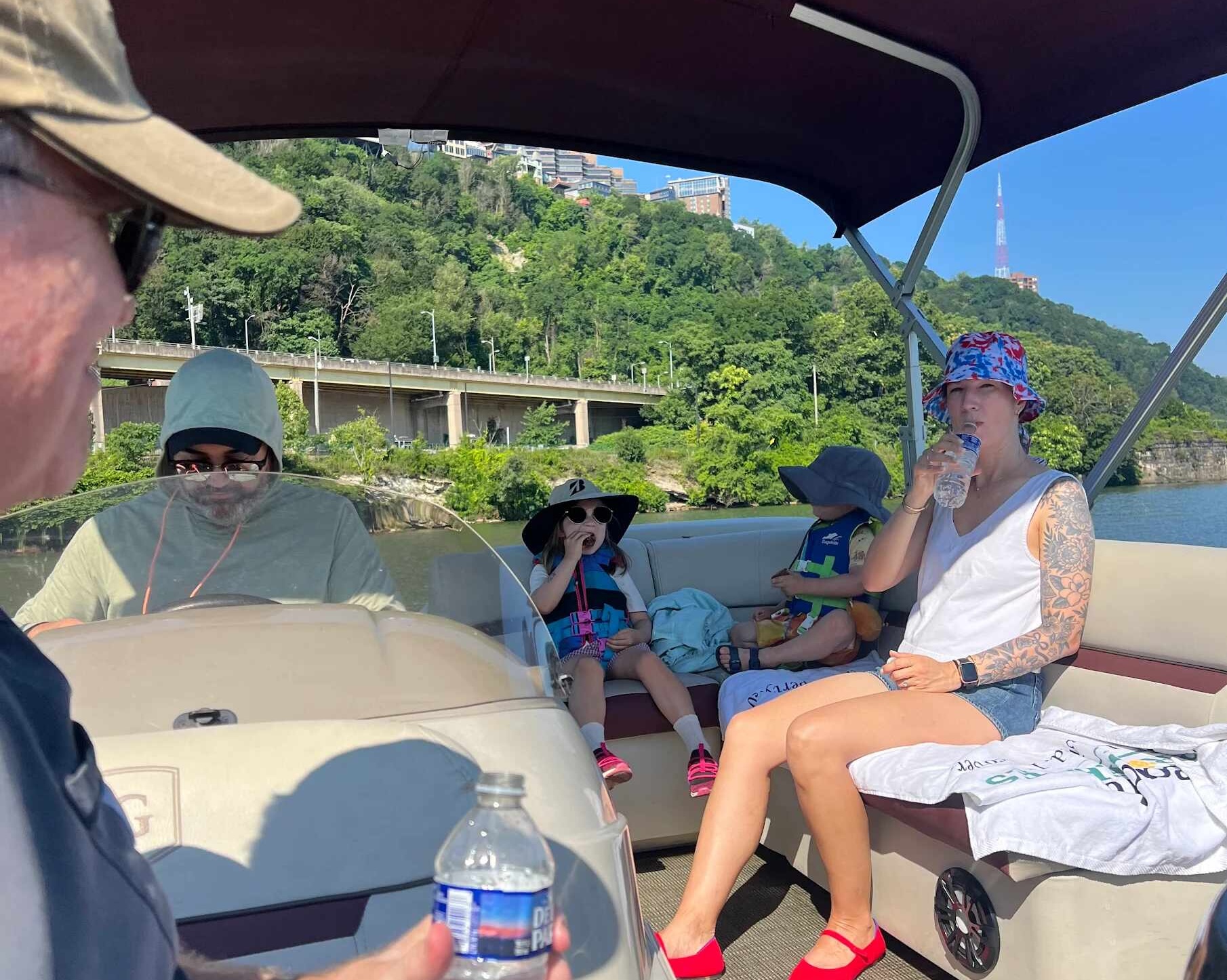 A family on a pontoon with the driver at the helm, two small kids in life jackets and sun hats, a woman drinking water, and Mount Washington behind.