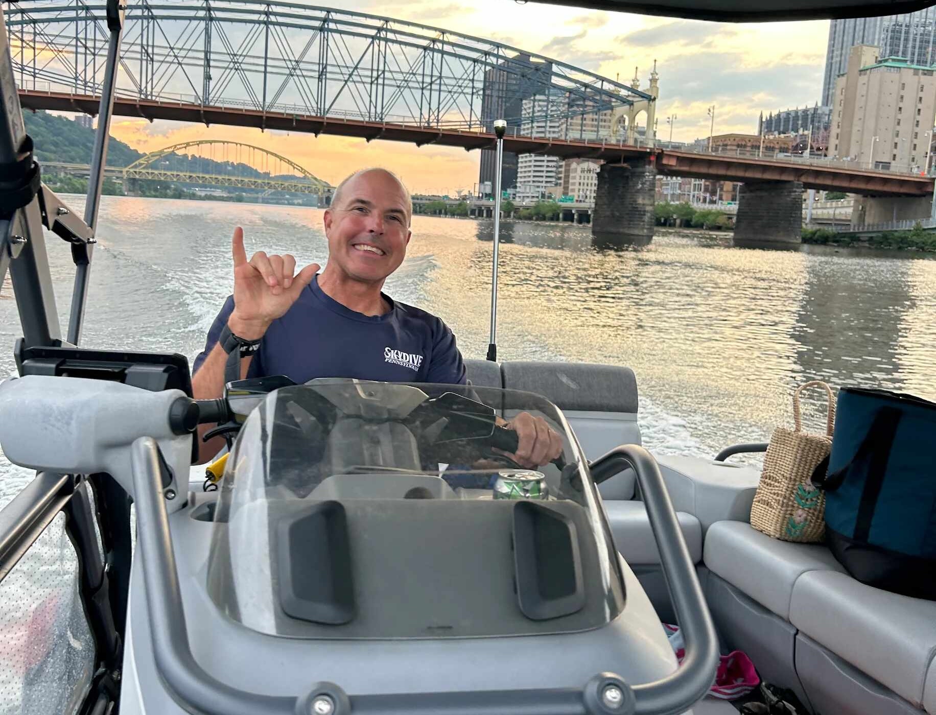 A smiling man making a hang loose sign at the helm of a Sea-Doo Switch, with a steel truss bridge and downtown buildings behind in golden light.