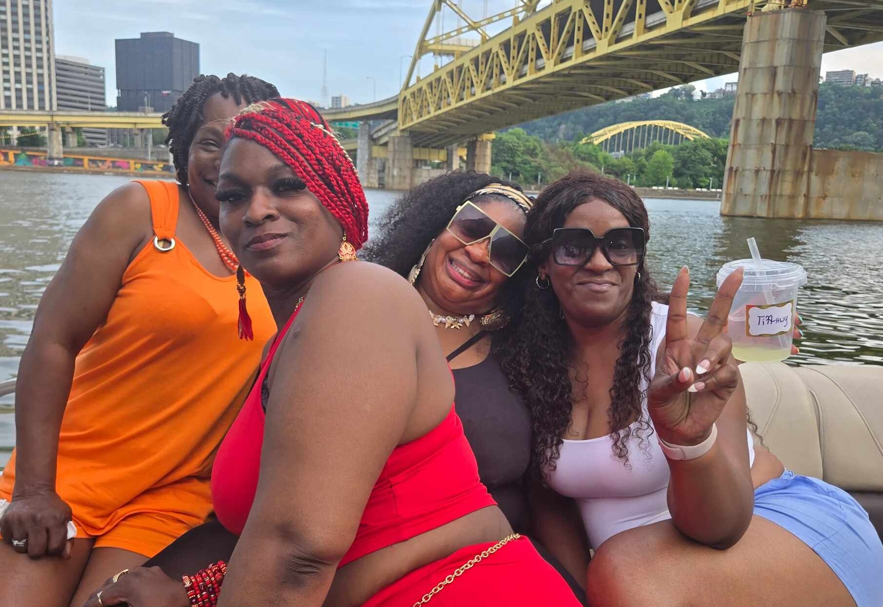 Four women smiling on a pontoon beneath a yellow double deck bridge, with another arch bridge and downtown buildings behind.