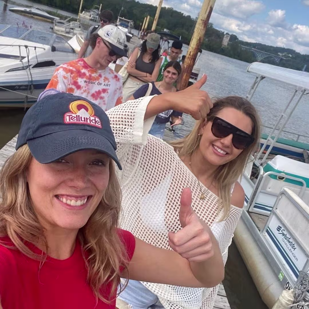 Two smiling women taking a selfie on the dock while friends board a Sweetwater pontoon behind them, with the river and a bridge in the background