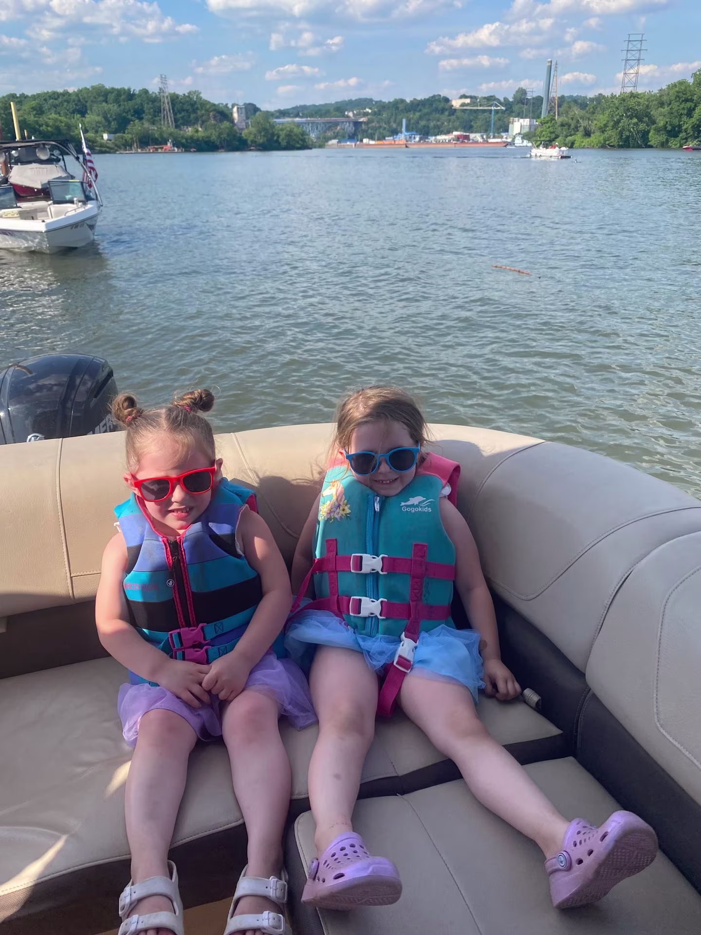 Two little girls in life jackets and sunglasses sitting on a pontoon bench on the river
