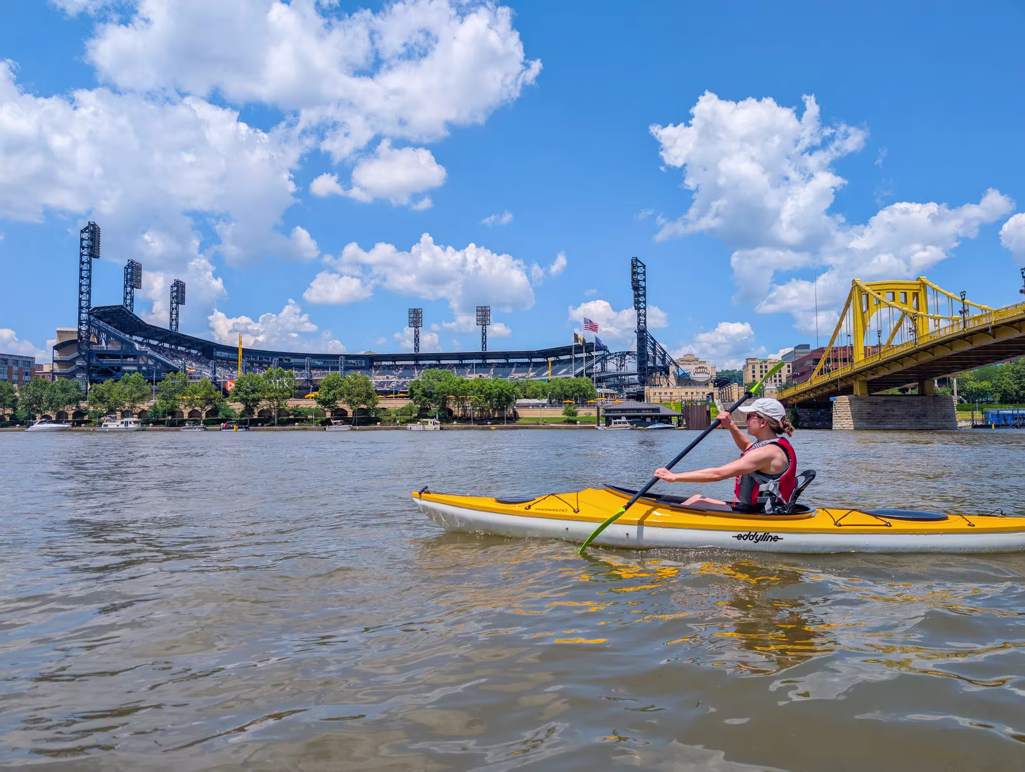 A woman in a life jacket paddling a yellow kayak on the river in front of PNC Park and a yellow bridge