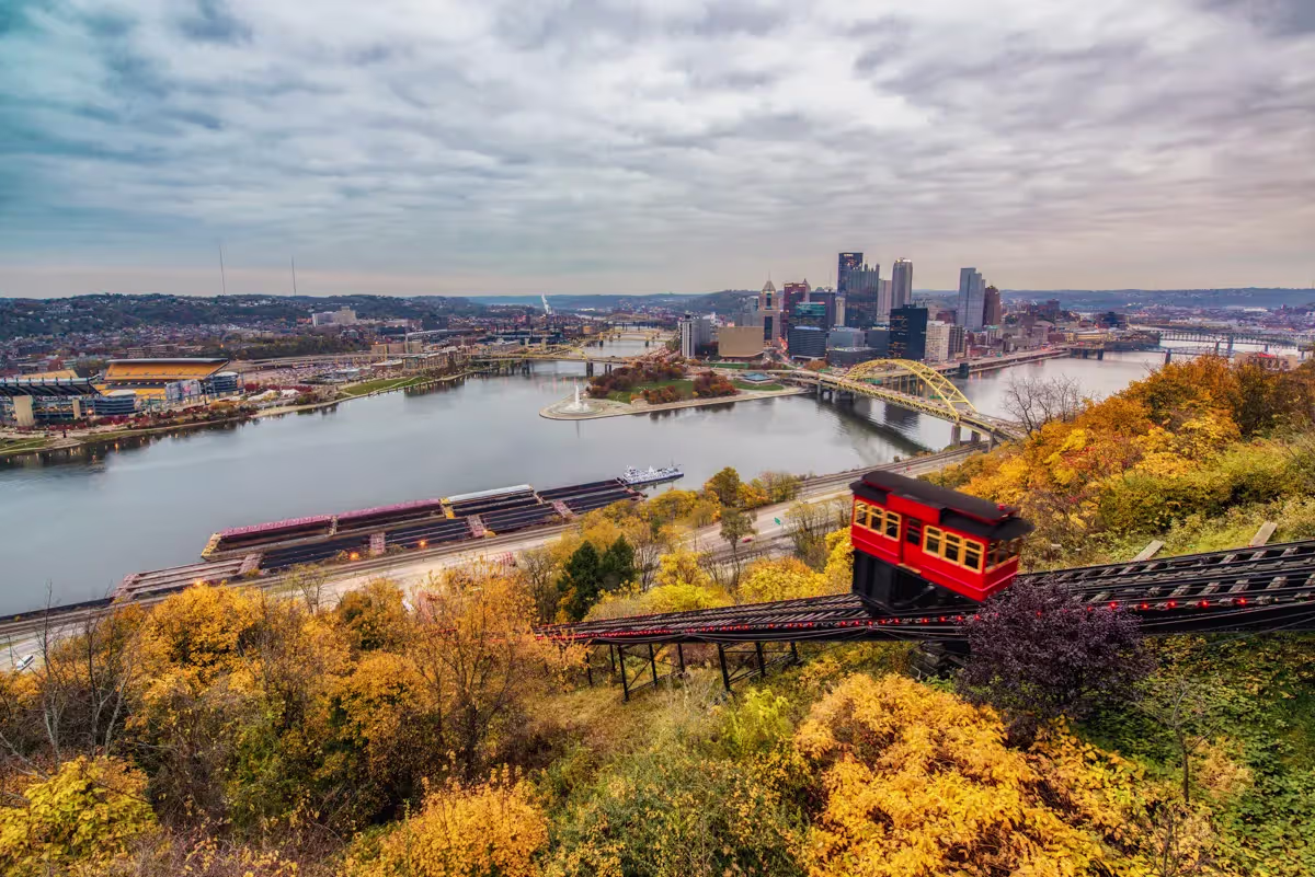 Red Duquesne Incline car on the hillside above yellow fall trees, with the Point, Fort Pitt Bridge and downtown skyline below