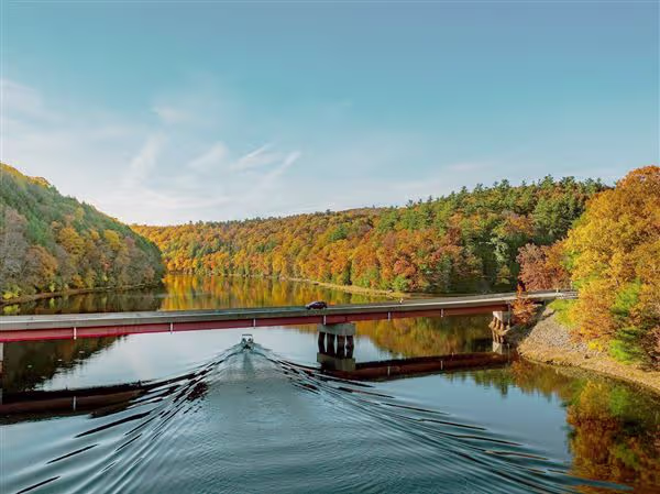 Boat traveling beneath a bridge on a tree-lined river surrounded by fall foliage