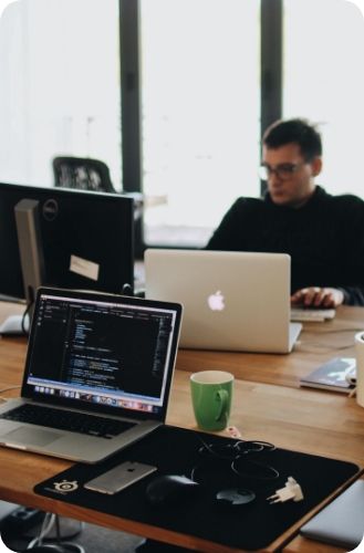 A person working on a laptop at a desk with another laptop displaying code and a green mug nearby.