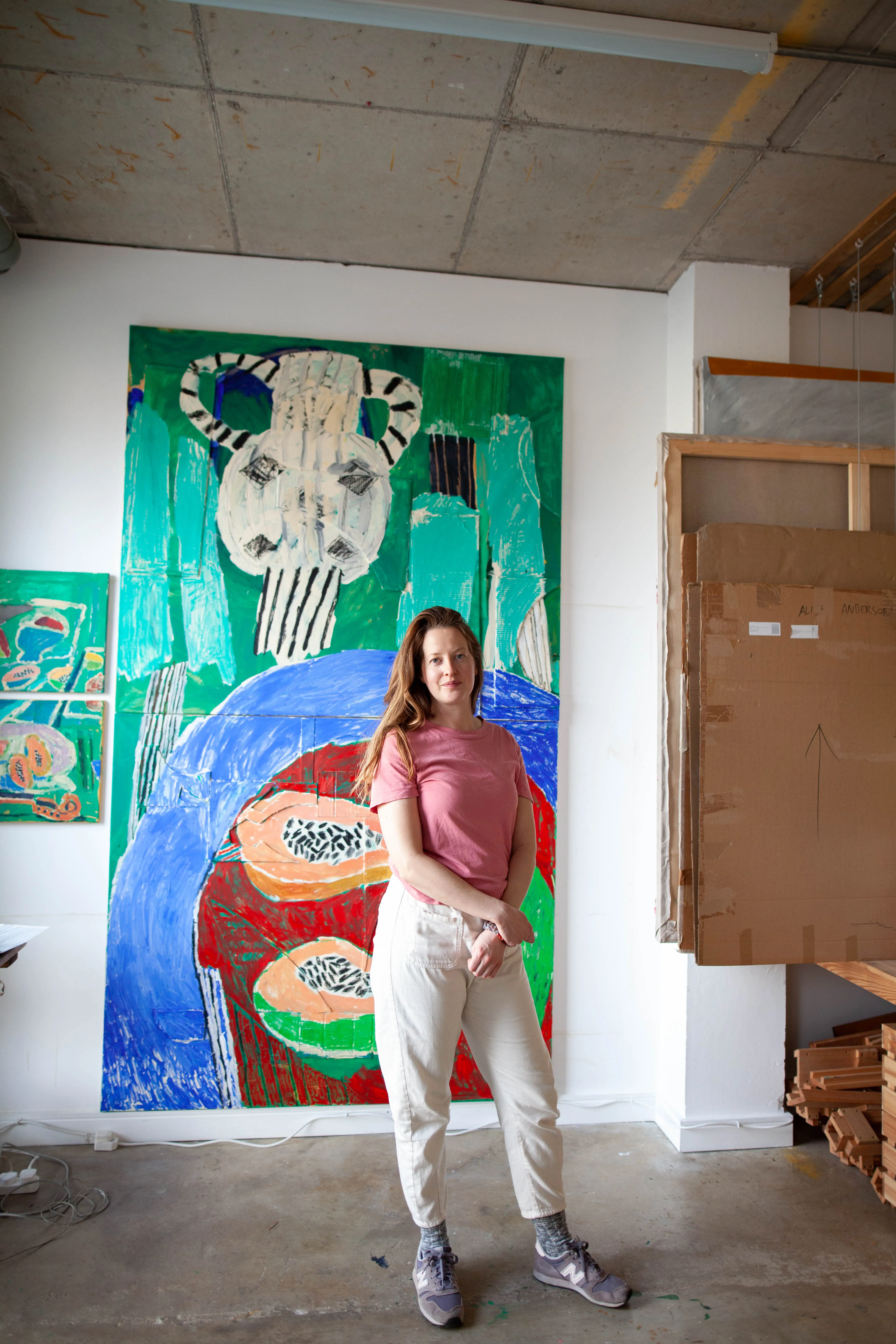 Young woman in pink t-shirt and white pants standing in front of large colorful abstract paintings in an artist's studio.