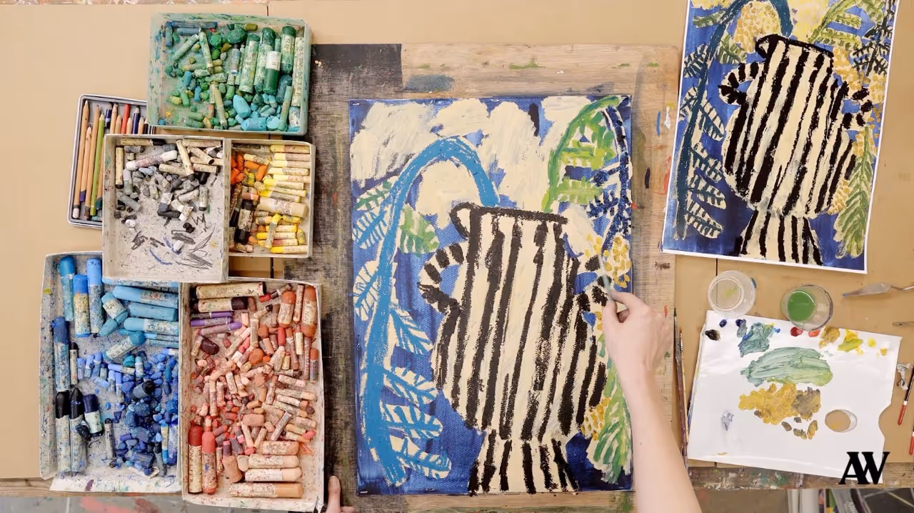 Floor covered with open sketchbooks showing colorful paintings, a cloth with black shapes, paint palettes, brushes, scissors, and other art supplies scattered around.