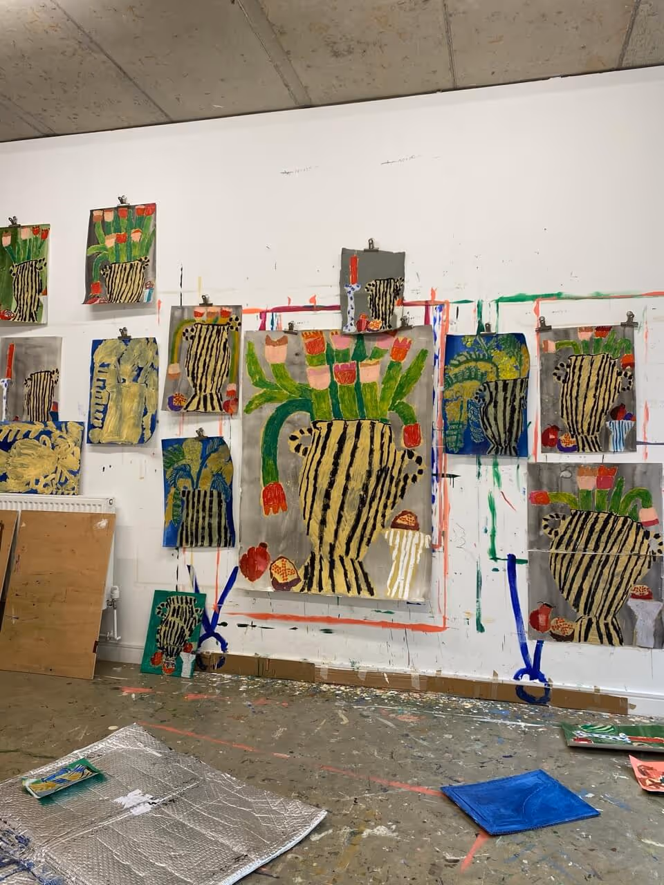 Art studio wall displaying multiple colorful paintings of striped vases with flowers, with paint splatters on the floor.