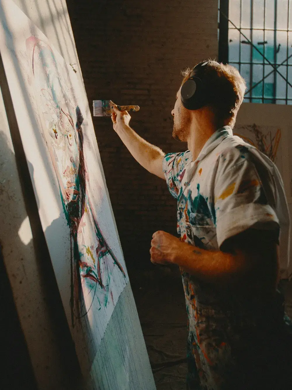 Artist wearing headphones and a paint-splattered shirt painting an abstract portrait on a large canvas in a studio with industrial windows.