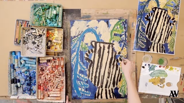 Floor covered with open sketchbooks showing colorful paintings, a cloth with black shapes, paint palettes, brushes, scissors, and other art supplies scattered around.