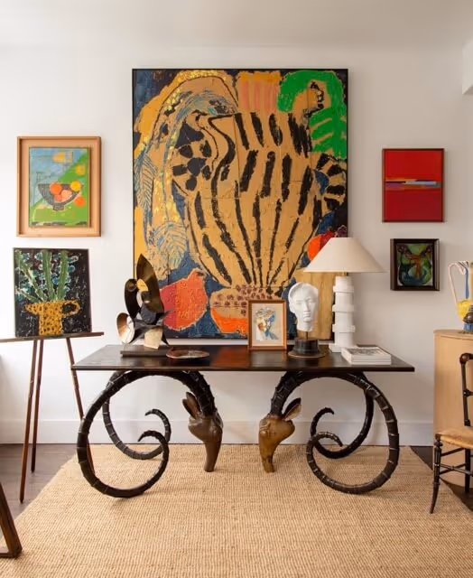Art-filled room with a black table featuring ornate curled legs and ram head details, topped with a white bust, a lamp, and various art pieces against a wall with colorful modern paintings.