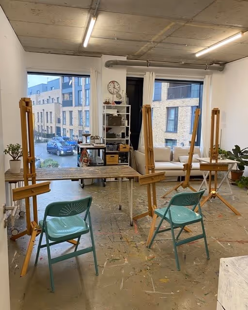 Art studio with wooden easels, two turquoise chairs, a worktable, and a window showing modern apartment buildings outside.