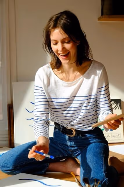 Smiling woman sitting cross-legged on the floor, holding a paintbrush and palette, in a sunlit room.
