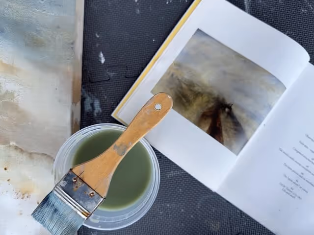 Paintbrush resting on a container of green paint next to an open book with a blurry image and text.