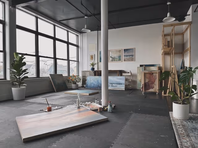 Bright art studio with large windows, several landscape paintings leaning against walls, potted plants, and art supplies on a black floor.