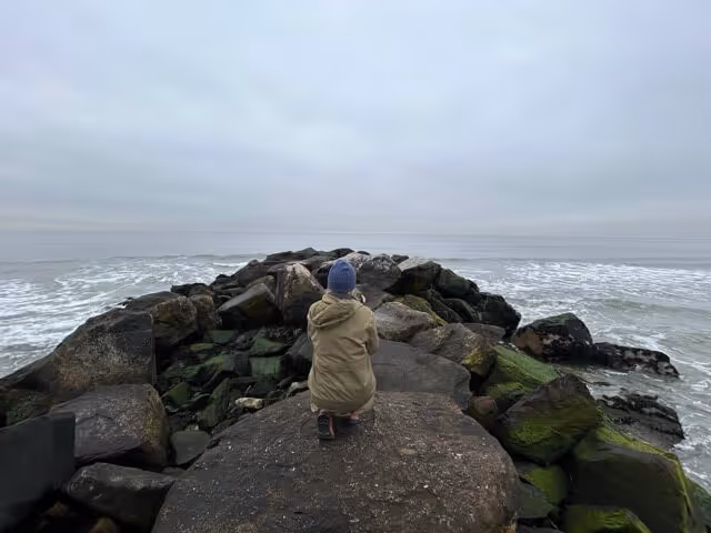 Person wearing a beige jacket and blue knit hat crouching on a jetty of large moss-covered rocks extending into the ocean under a cloudy sky.