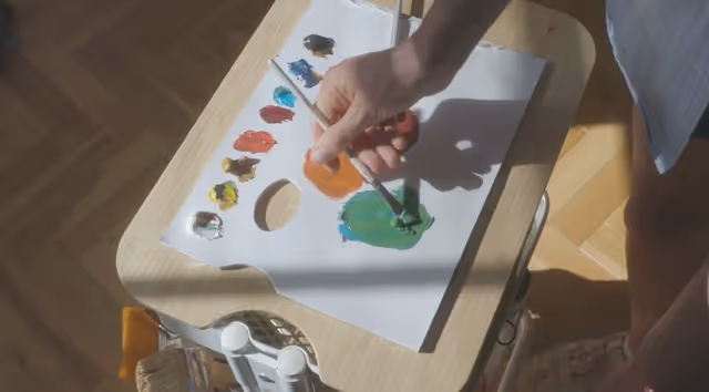 Person mixing green paint with a brush on a palette filled with various paint colors on a wooden table.