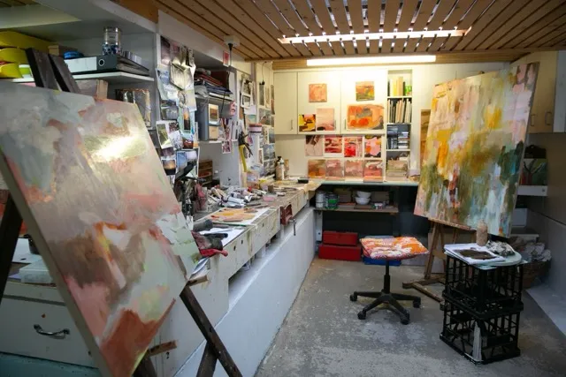 Artist studio with two large abstract paintings on easels, a swivel chair, and shelves filled with art supplies and small artworks on the walls.