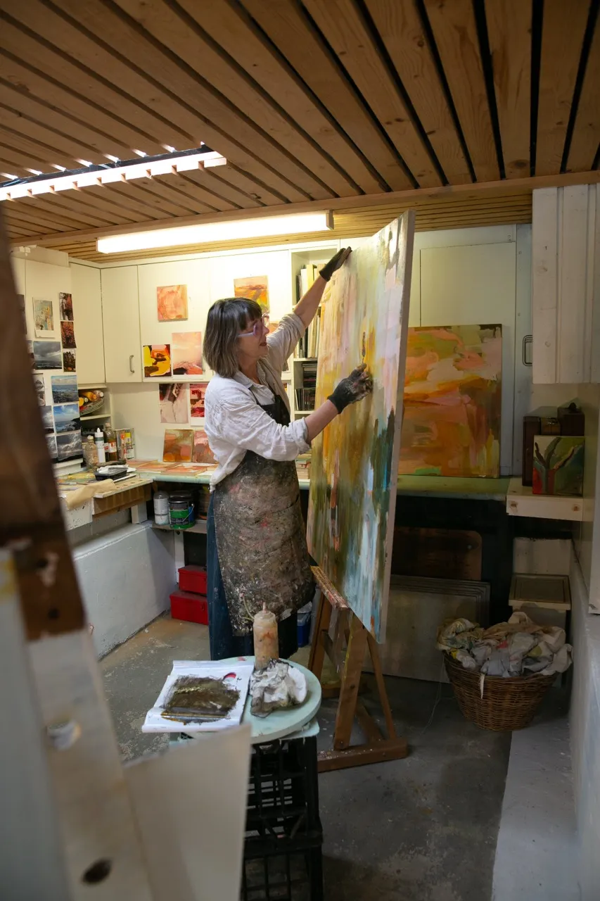 Artist wearing gloves and apron working on a large abstract painting in a well-lit studio filled with art supplies and other paintings.