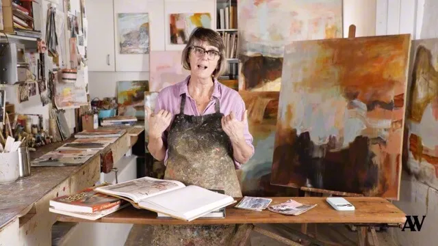Woman wearing glasses and a paint-stained apron speaking in an art studio with canvases and art supplies around her.