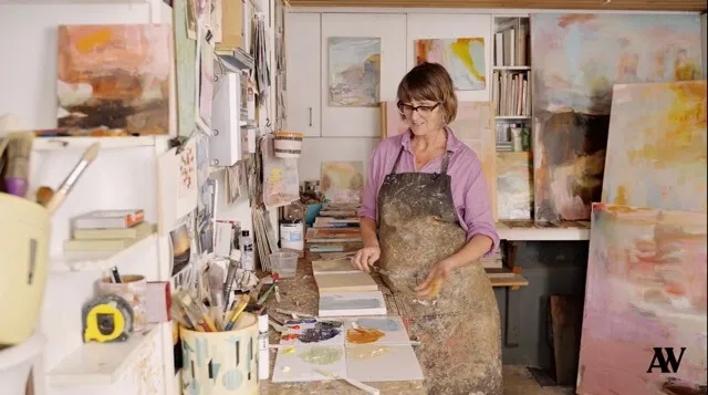 Woman artist in a paint-stained apron working on multiple abstract paintings in a studio filled with art supplies and canvases.