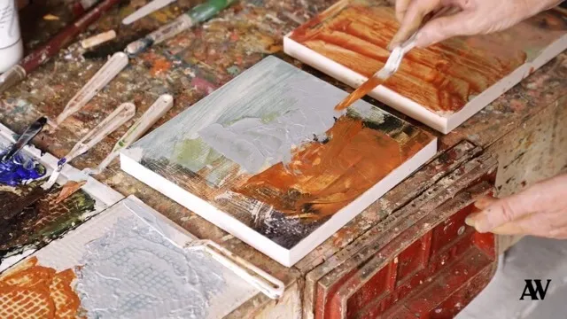Hands applying brown oil paint with a brush on a canvas among other painted canvases on a workbench.