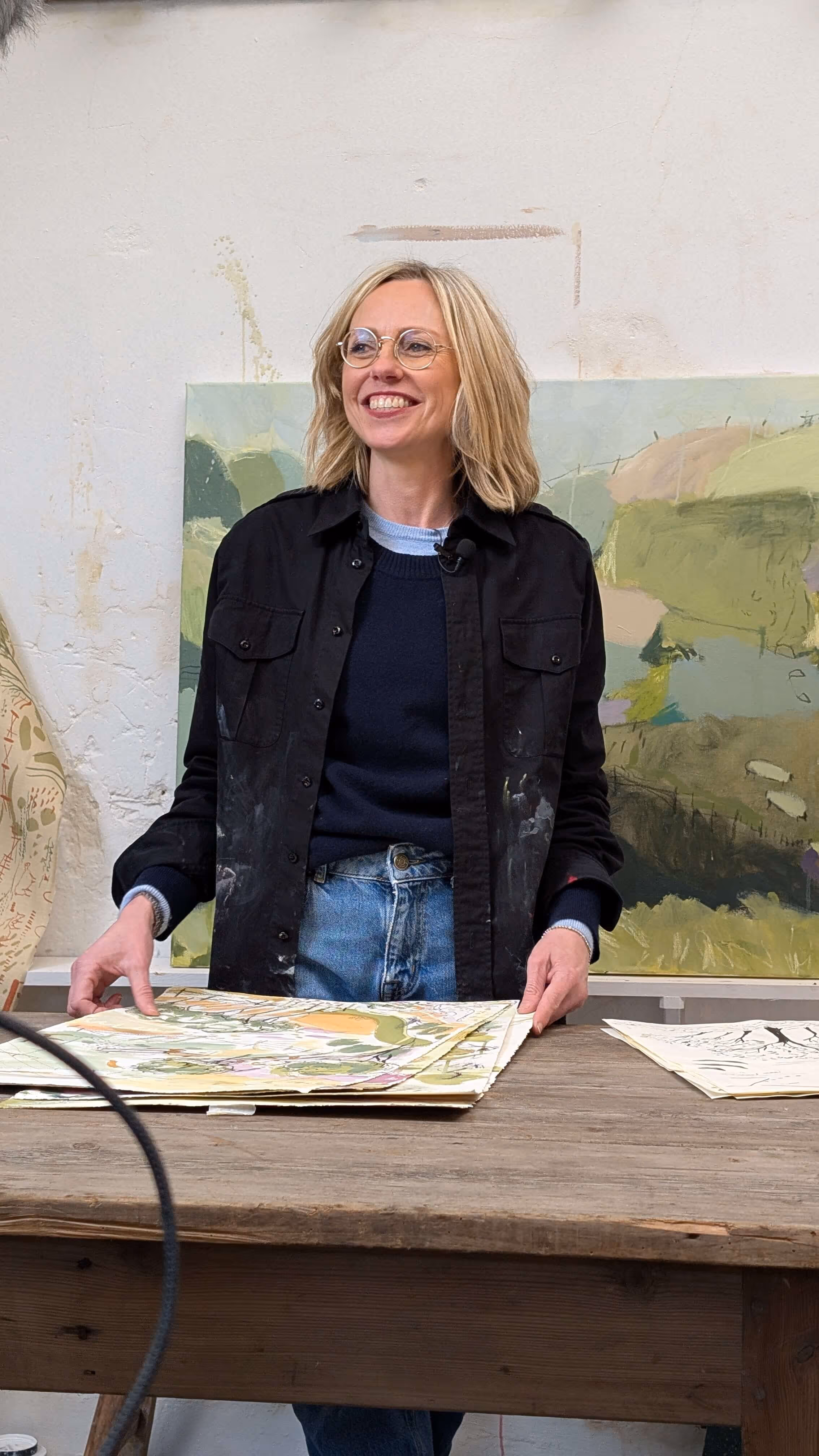 Smiling woman with glasses stands behind table with paintings and a landscape artwork in background.