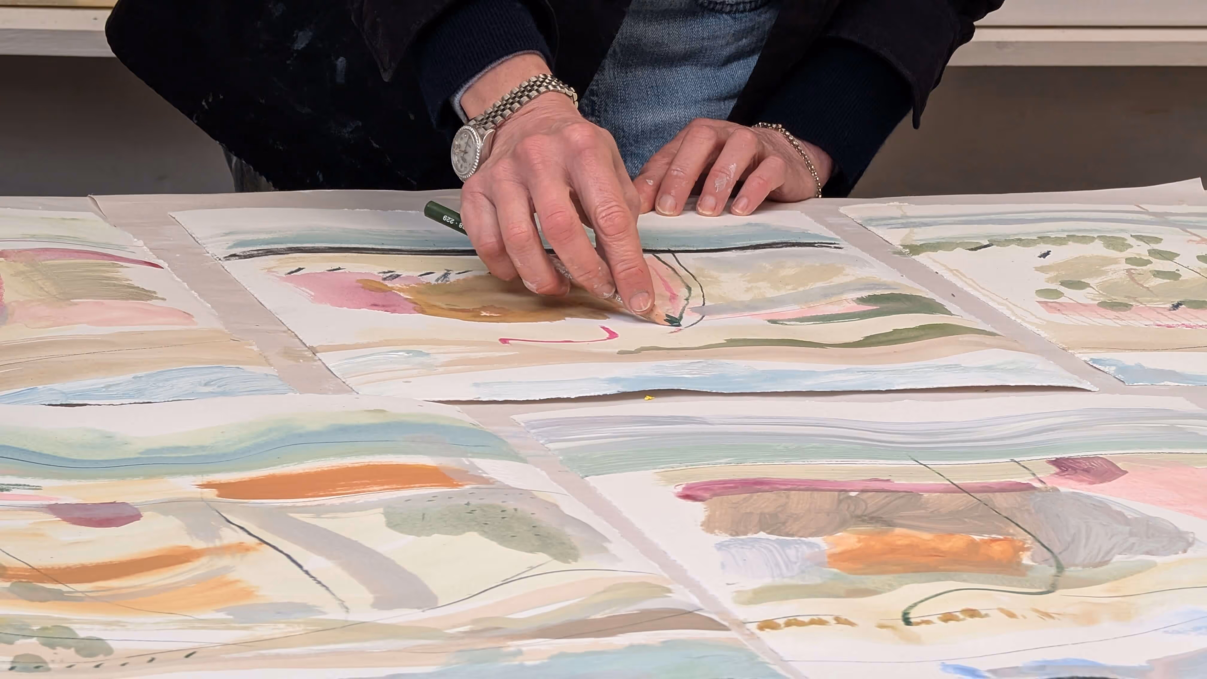 Person drawing with green pencil on colorful abstract paintings spread on a table.