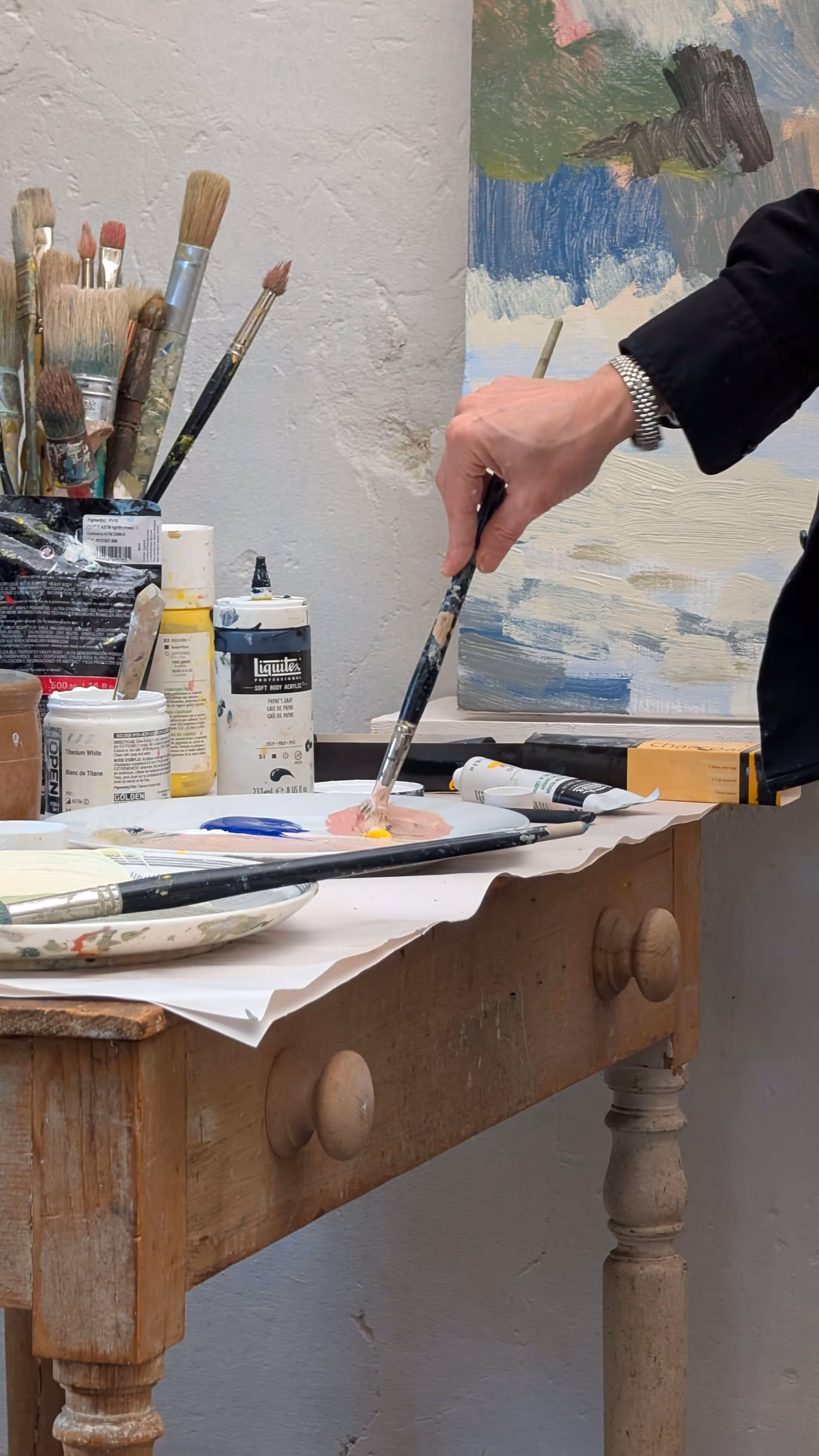 Hand mixing paint with a brush on a palette next to jars and brushes on a wooden table.