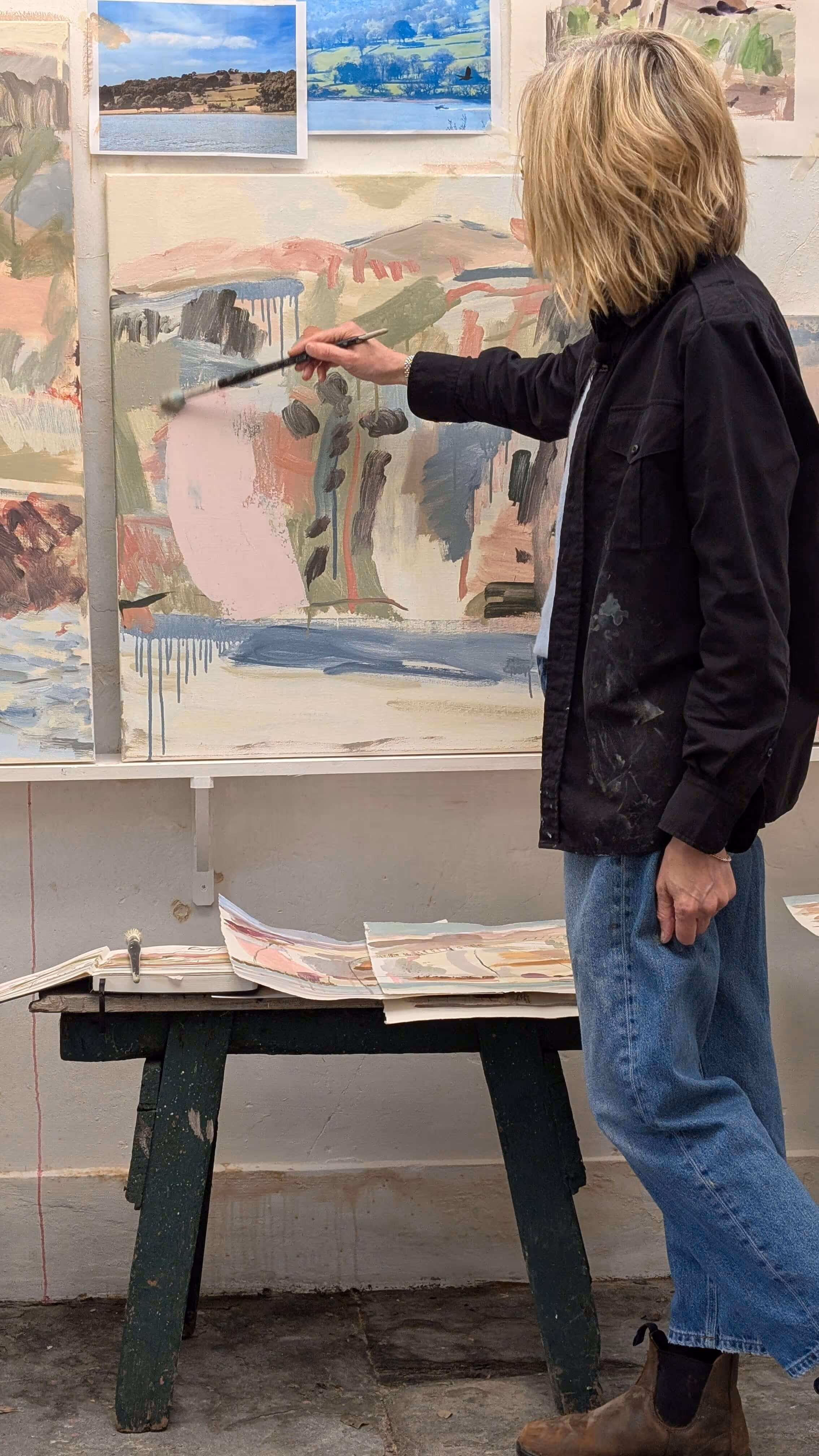 Artist in black shirt and jeans painting abstract canvas in studio with landscape photos pinned up.
