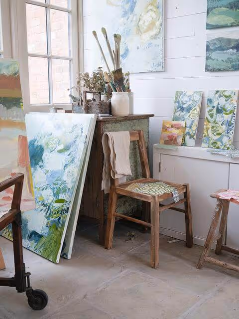 A bright artist studio with abstract floral paintings, brushes in jars, and wooden chairs.