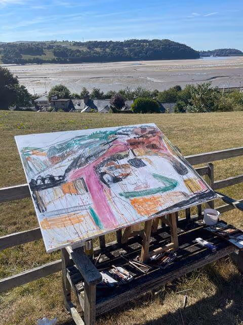Abstract painting with pink, orange, and green on easel outdoors by a wooden fence and shoreline.