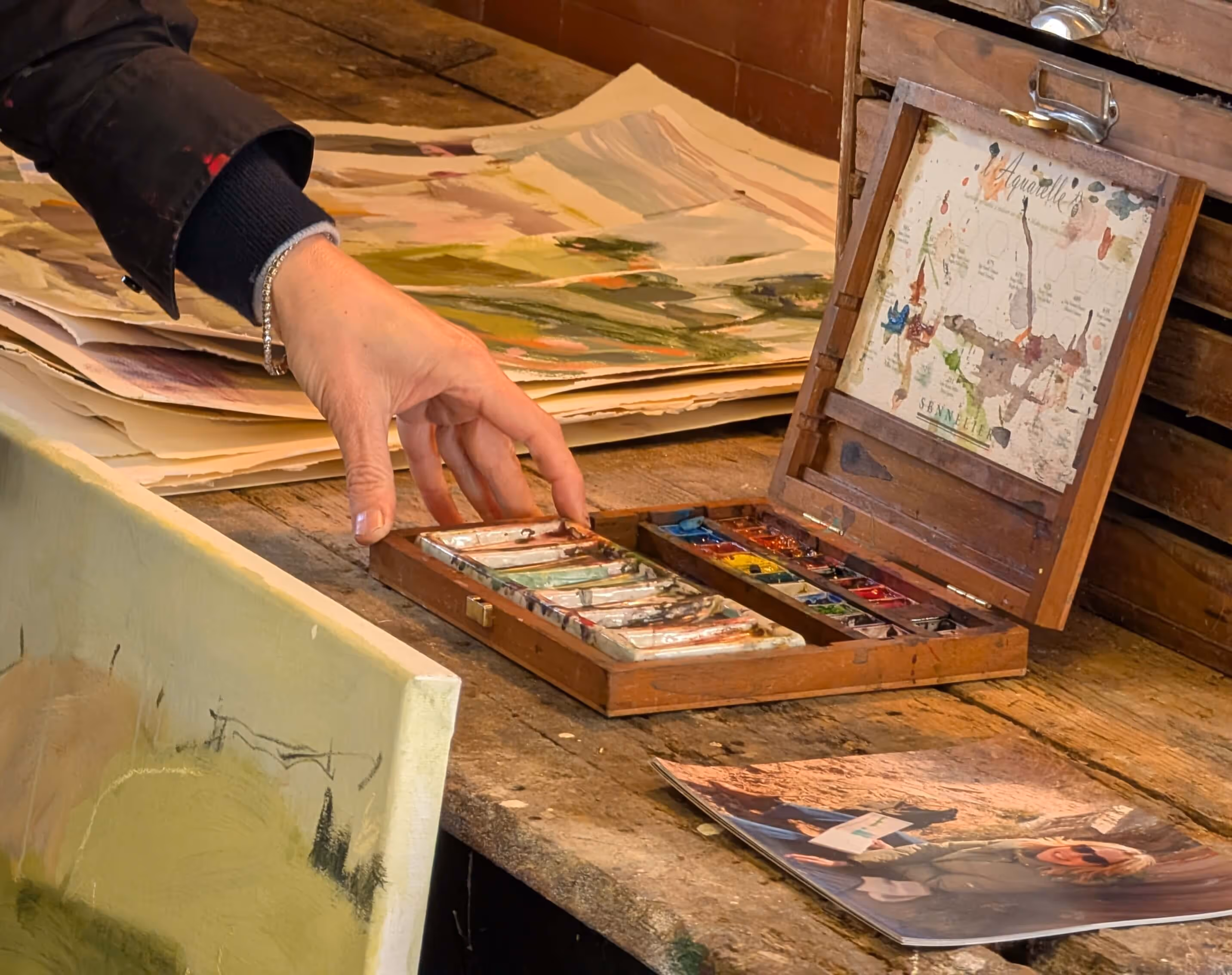 Hand reaching for watercolor paint palette on a wooden table with paper and canvas nearby.