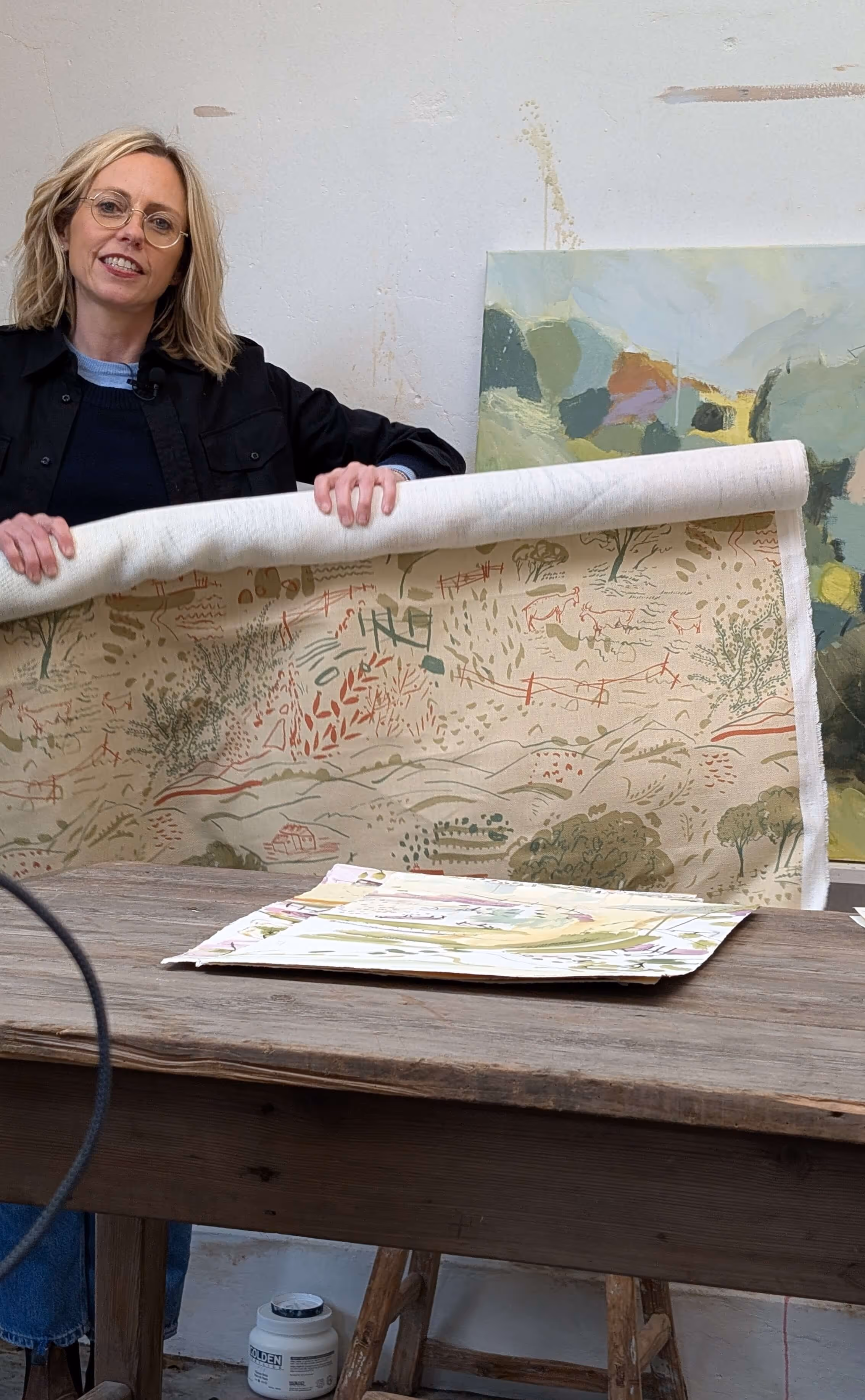 Woman with glasses holds a roll of fabric printed with nature-inspired patterns at a wooden table.