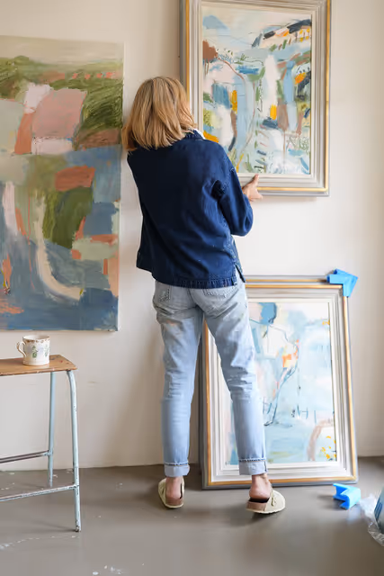 Woman hanging framed abstract painting on white wall next to another painting and a cup on a stool.