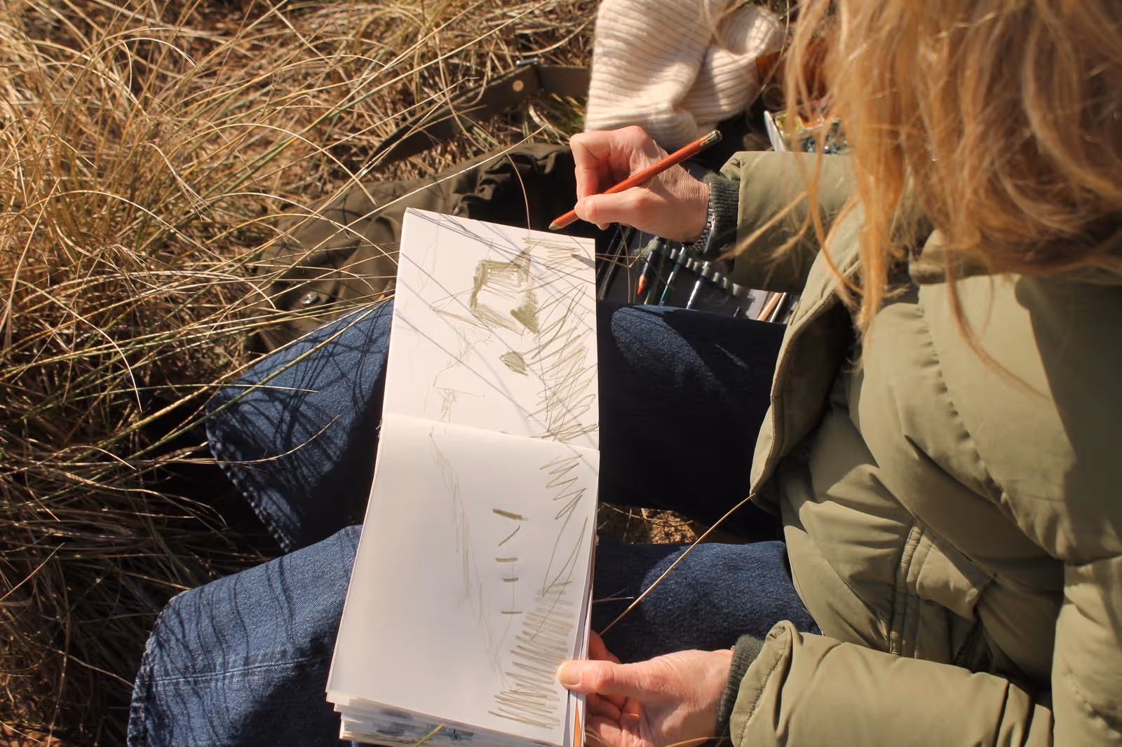 Person sitting outdoors sketching a landscape in a notebook, surrounded by dry grass.