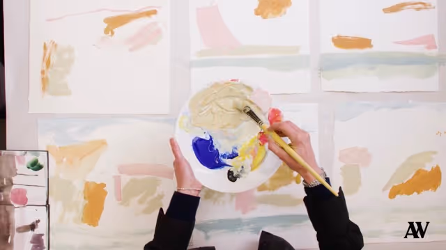Hands mixing beige paint on a palette above abstract paintings with pastel colors on a table.