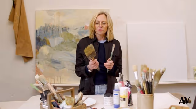 Artist holding two brushes in a studio with paint supplies and canvases behind her.